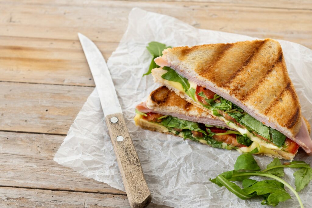 Sandwich grillé garni de jambon et de roquette posé sur du papier, à côté d'un couteau, sur une table en bois