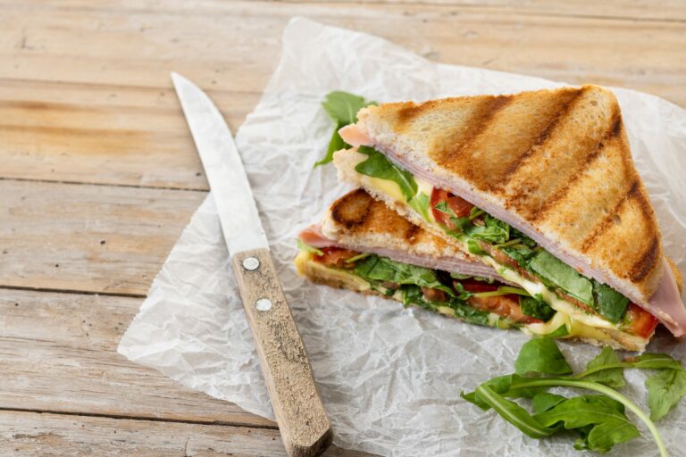 Sandwich grillé garni de jambon et de roquette posé sur du papier, à côté d'un couteau, sur une table en bois