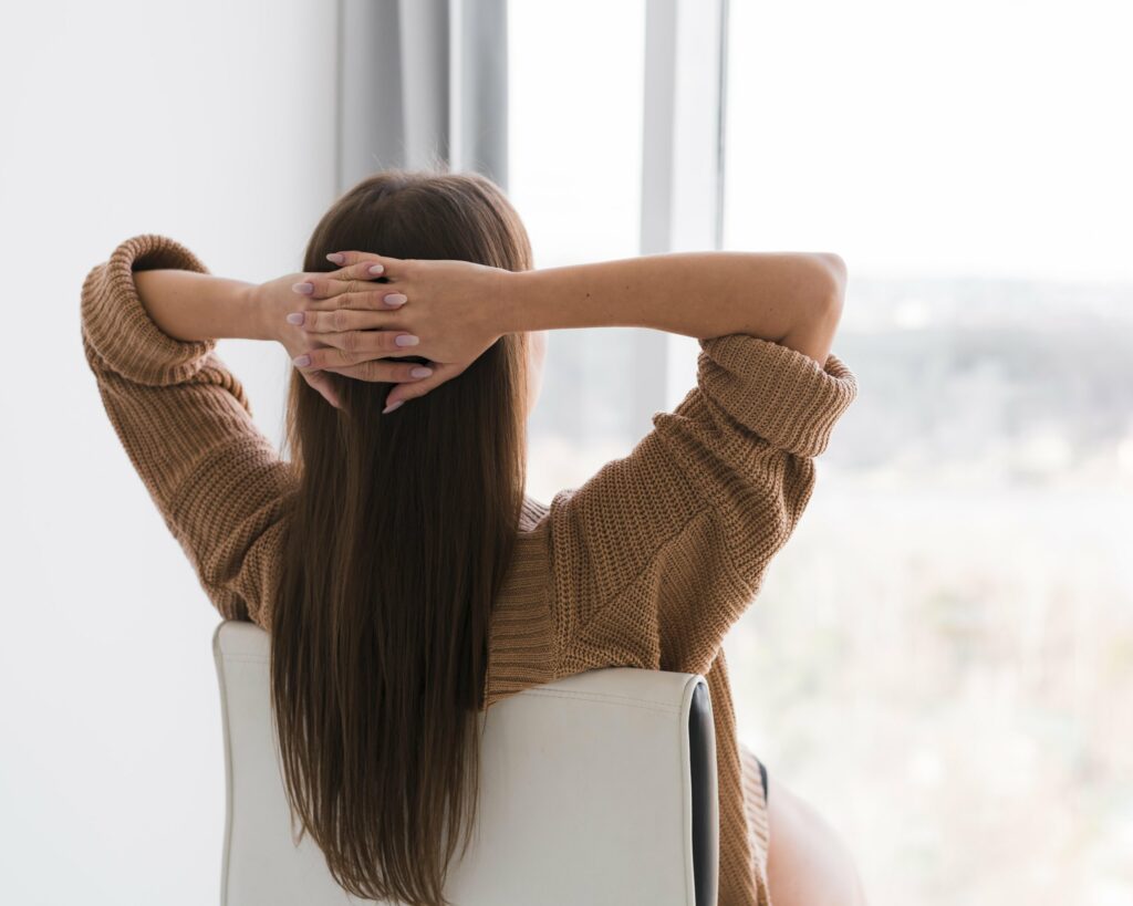 Femme de dos, les mains derrière la tête, face à une fenêtre