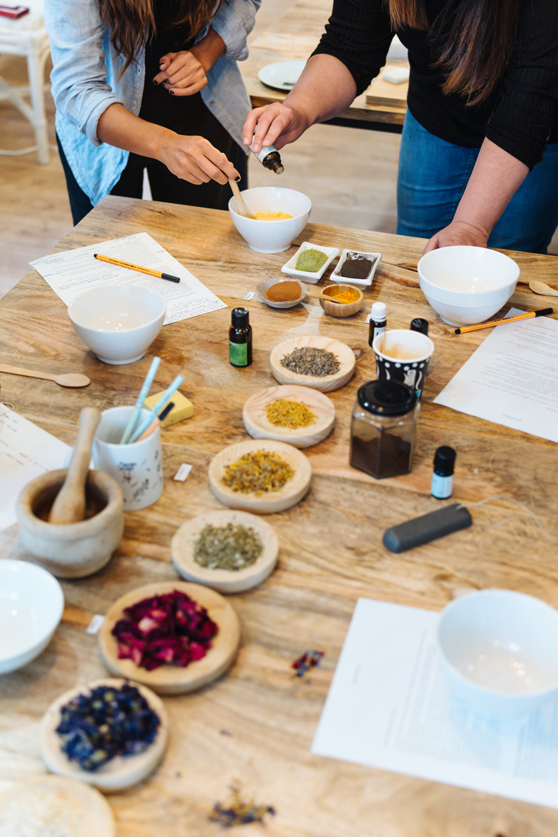 Table d’atelier en bois garnie de fleurs séchées, d’épices et de flacons d’huiles essentielles, deux participantes à l’œuvre