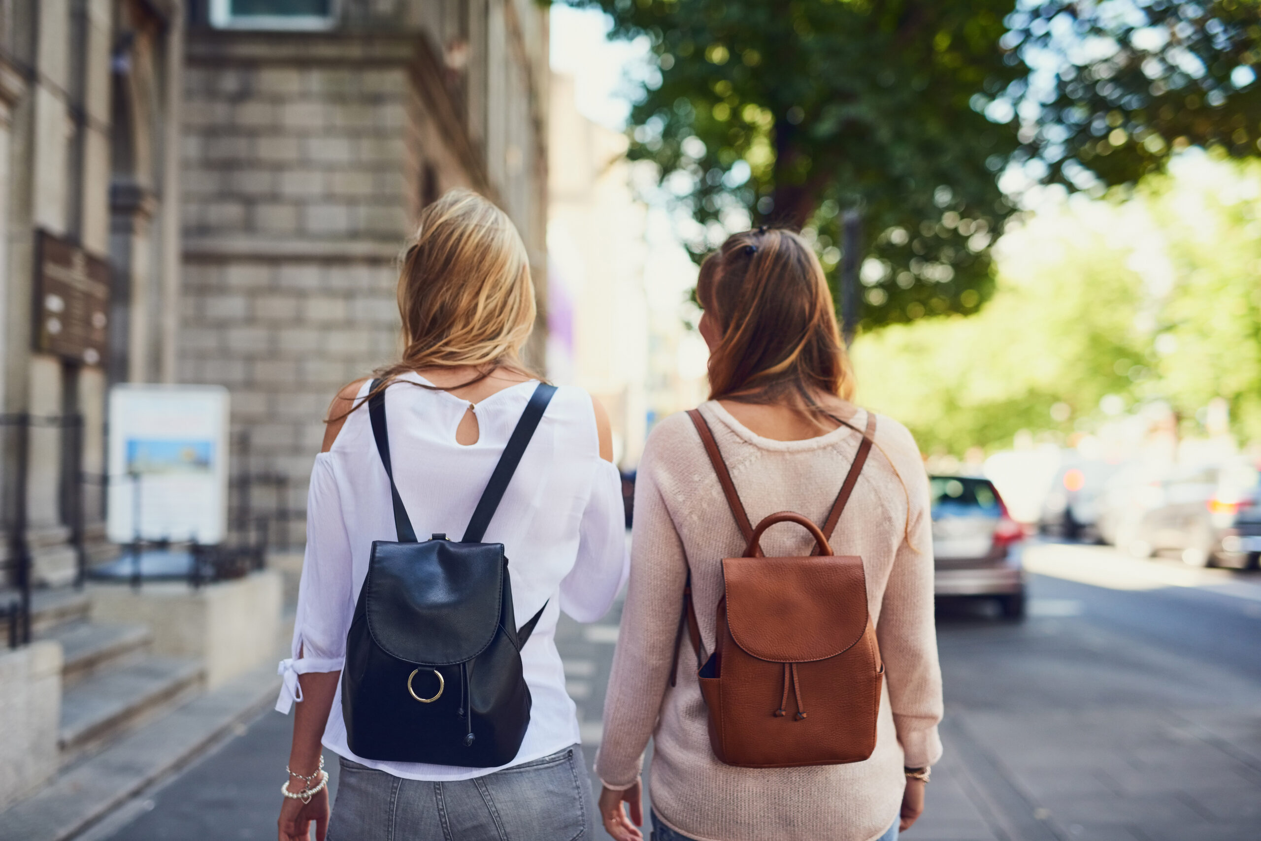 Deux femmes vues de dos, sacs à dos en cuir noir et camel, se promènent côte à côte dans une rue de ville arborée