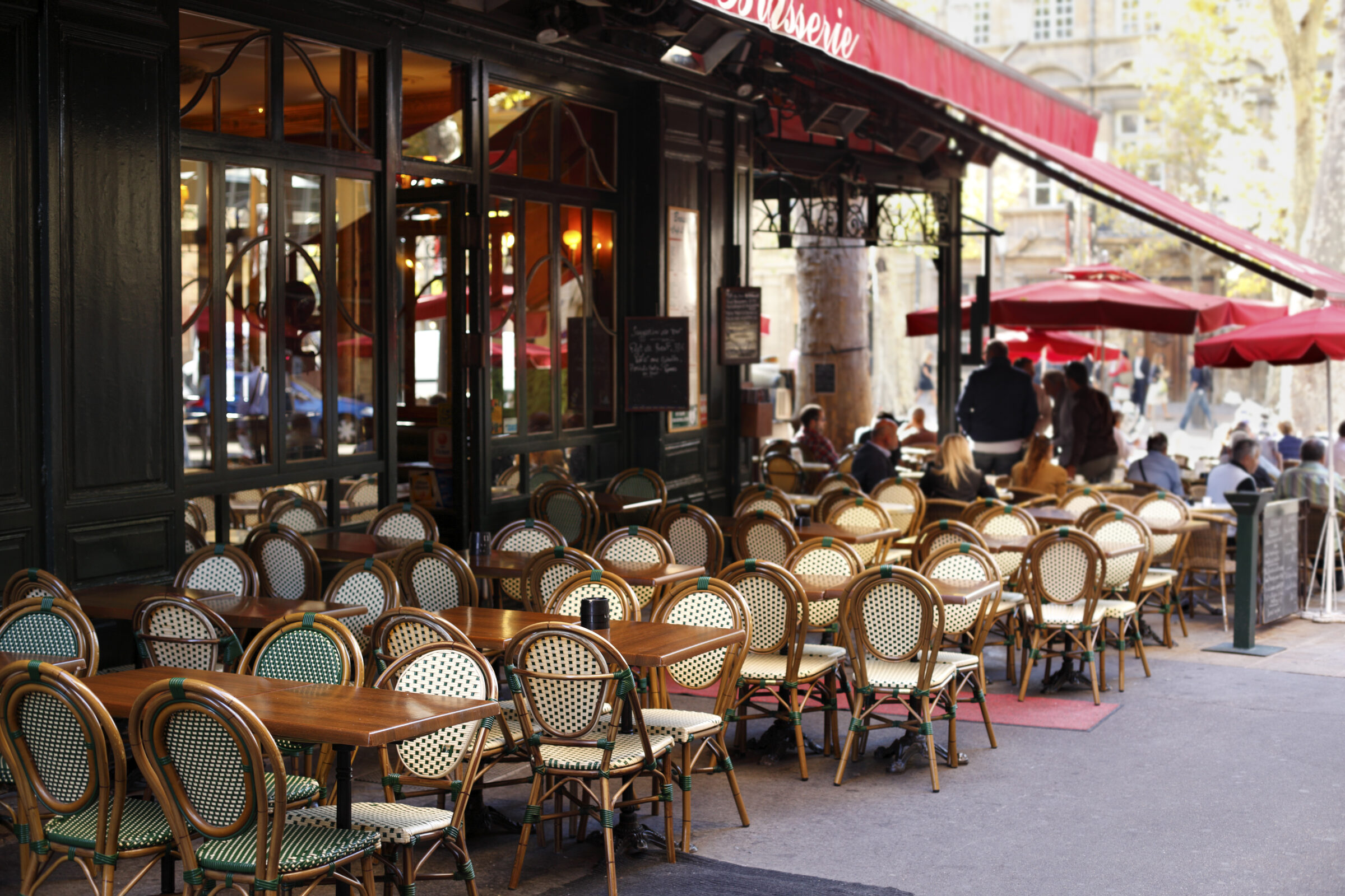 Terrasse d’une brasserie parisienne au soleil, chaises en rotin et clients attablés sous les auvents rouges