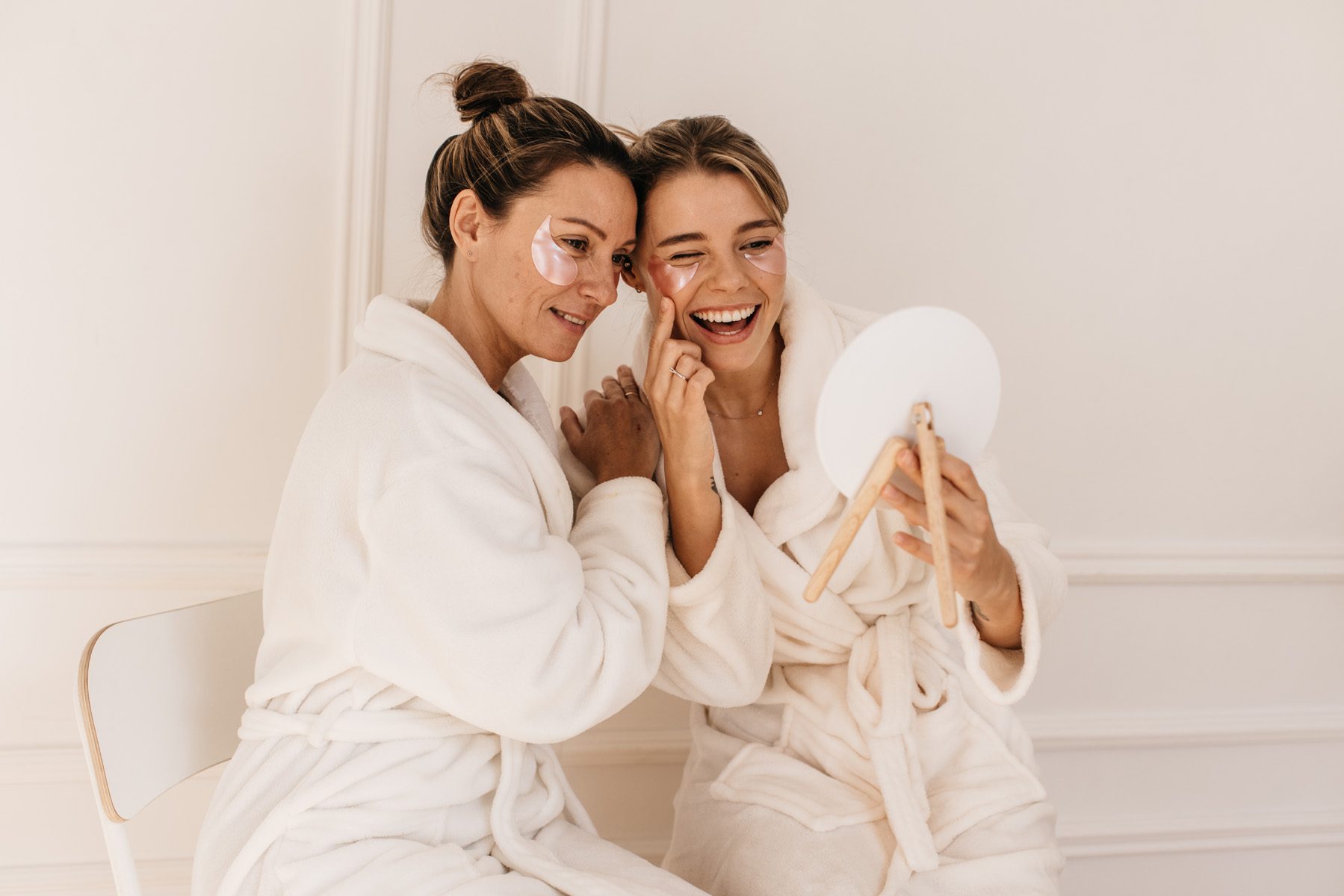 Une mère et sa fille en peignoir blanc, patchs sous les yeux, riant ensemble devant un petit miroir