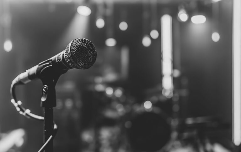 Microphone sur pied au premier plan d’une scène de concert éclairée par des projecteurs, en noir et blanc