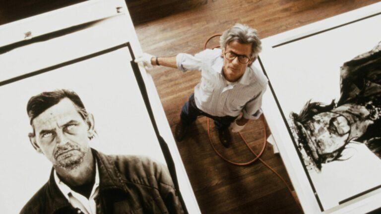 Homme debout dans un atelier au parquet, vu en plongée, entouré de grands tirages de portraits en noir et blanc, dans le documentaire Avedon