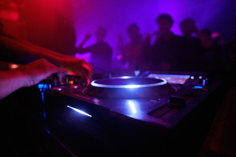 Platine de DJ éclairée au premier plan devant des danseurs baignés de lumière violette dans un club