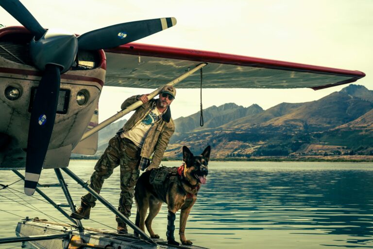 Un homme en tenue de camouflage et son chien sur le flotteur d'un hydravion, au bord d'un lac de montagne, dans Heart of the Beast