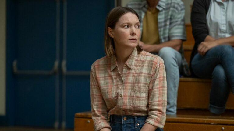 Une lycéenne en chemise à carreaux assise sur les gradins d’un gymnase, dans la série « Carrie »