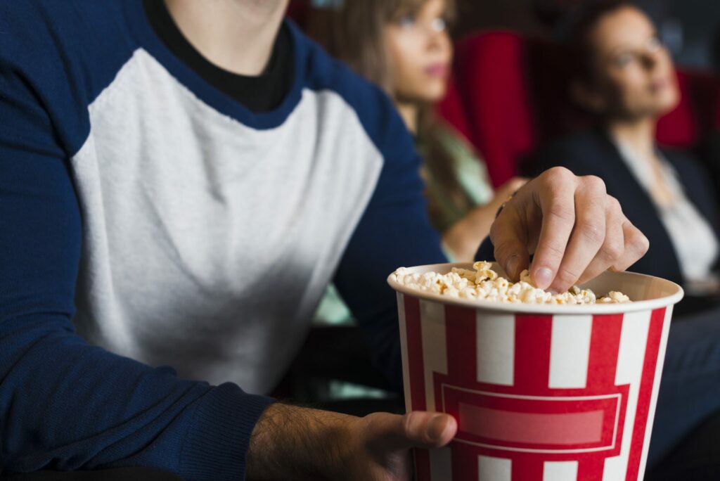 Spectateurs assis dans une salle de cinéma aux fauteuils rouges, un pot de pop-corn rayé rouge et blanc au premier plan