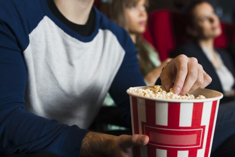 Spectateurs assis dans une salle de cinéma aux fauteuils rouges, un pot de pop-corn rayé rouge et blanc au premier plan