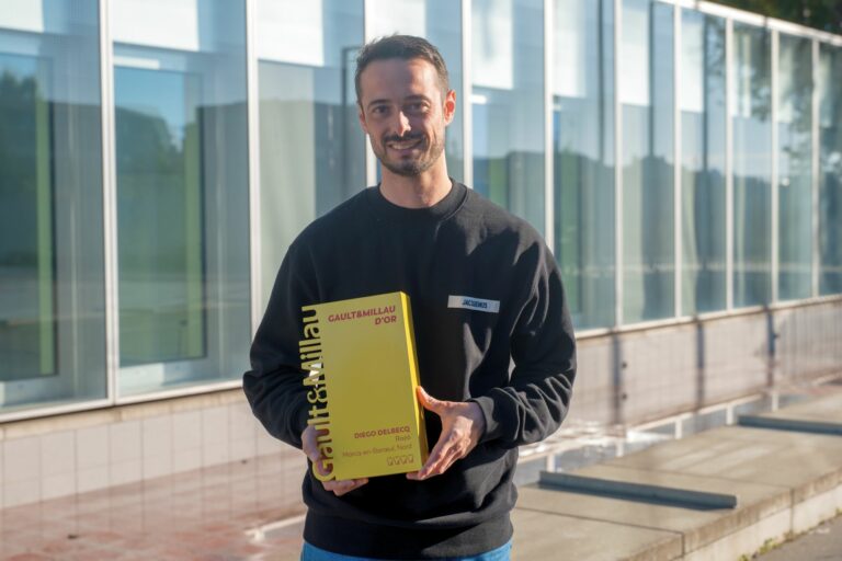 Le chef Diego Delbecq tenant le guide Gault&Millau devant un bâtiment vitré