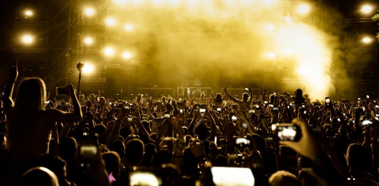 Foule de spectateurs vue de dos, téléphones levés, face à une scène de concert inondée de lumière dorée