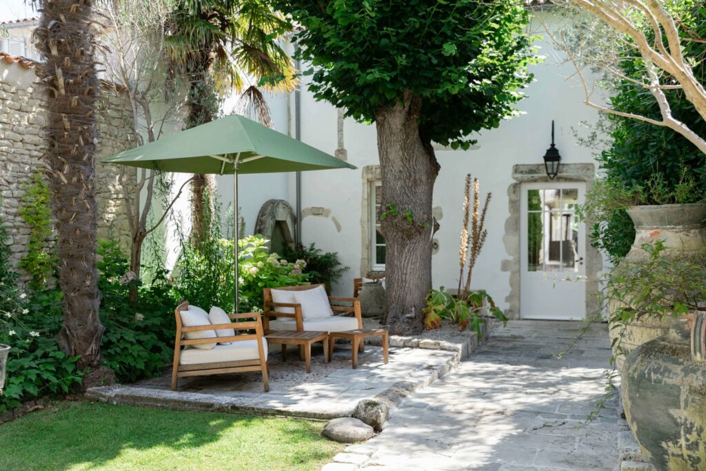 Terrasse de la Villa Clarisse sous les palmiers : fauteuils de bois, parasol vert et porte-fenêtre ouverte sur la maison