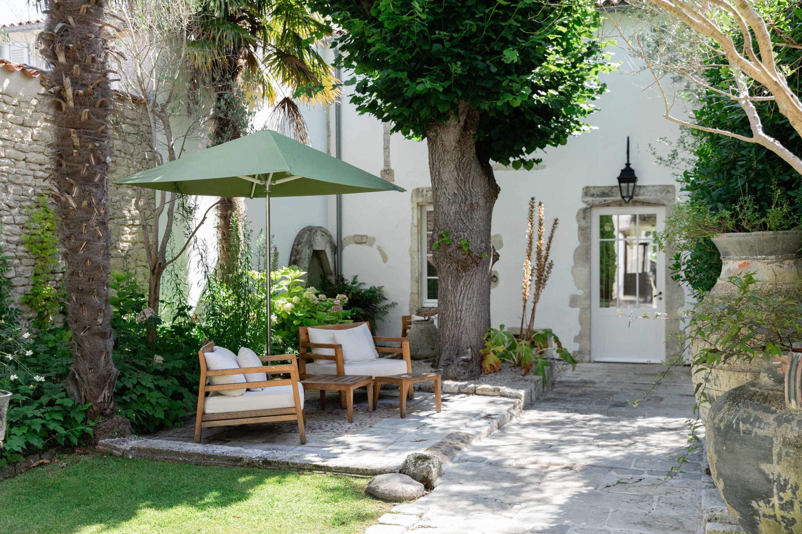 Terrasse de la Villa Clarisse sous les palmiers : fauteuils de bois, parasol vert et porte-fenêtre ouverte sur la maison