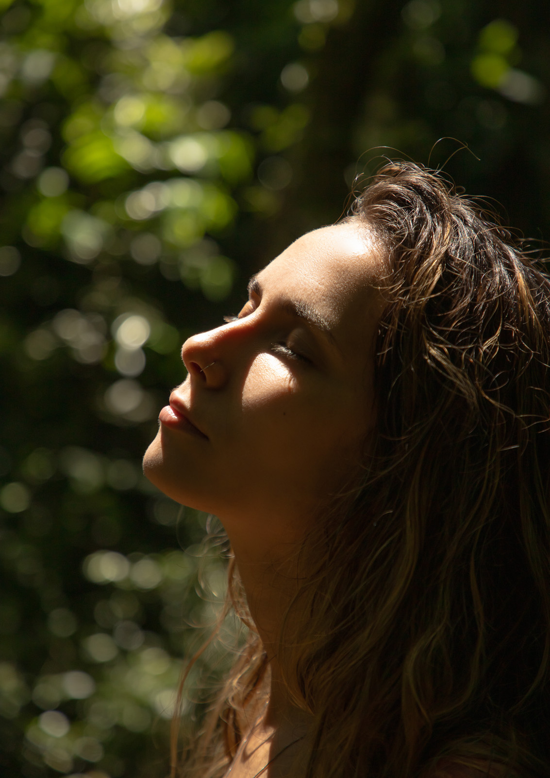 Femme les yeux fermés, visage levé vers la lumière du soleil, feuillage en fond