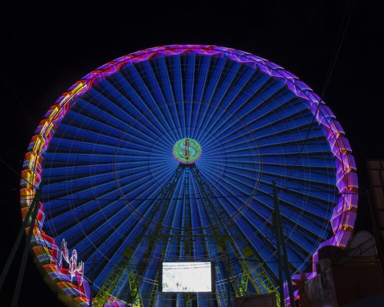 Grande roue de fête foraine illuminée de nuit, vue en contre-plongée, avec des rayons bleus et un pourtour rose, violet et orange sur un ciel noir