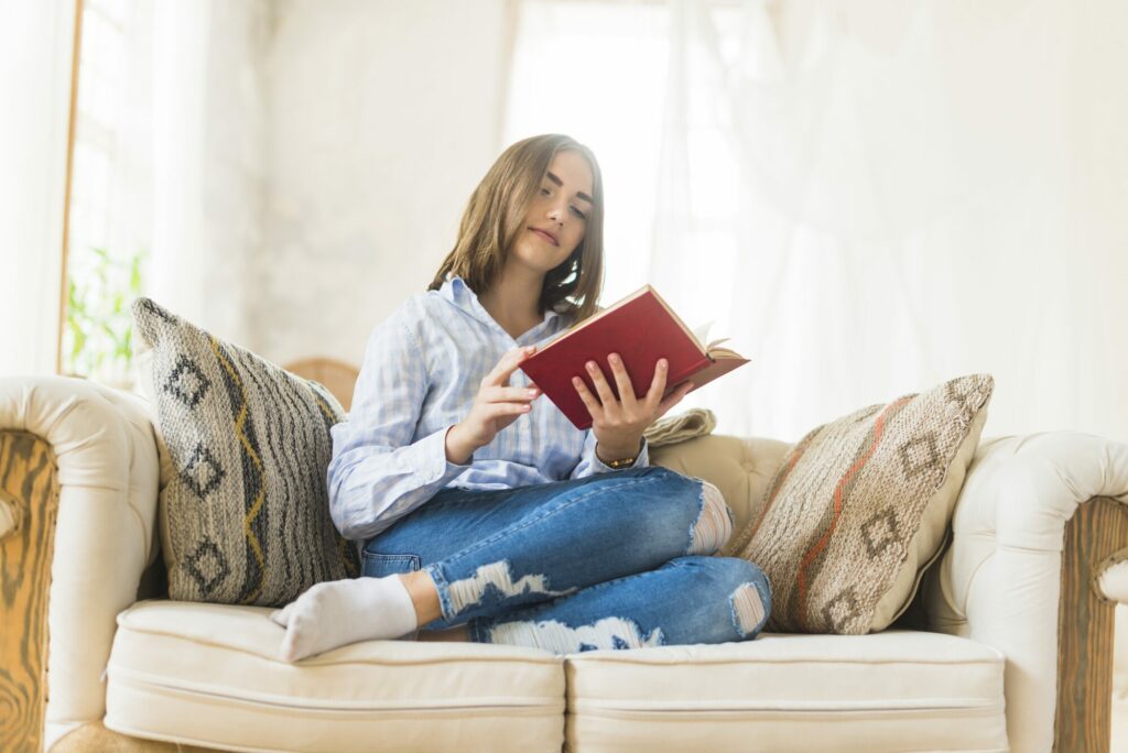 Jeune femme assise en tailleur sur un canapé blanc, lisant un livre à couverture rouge dans un salon lumineux