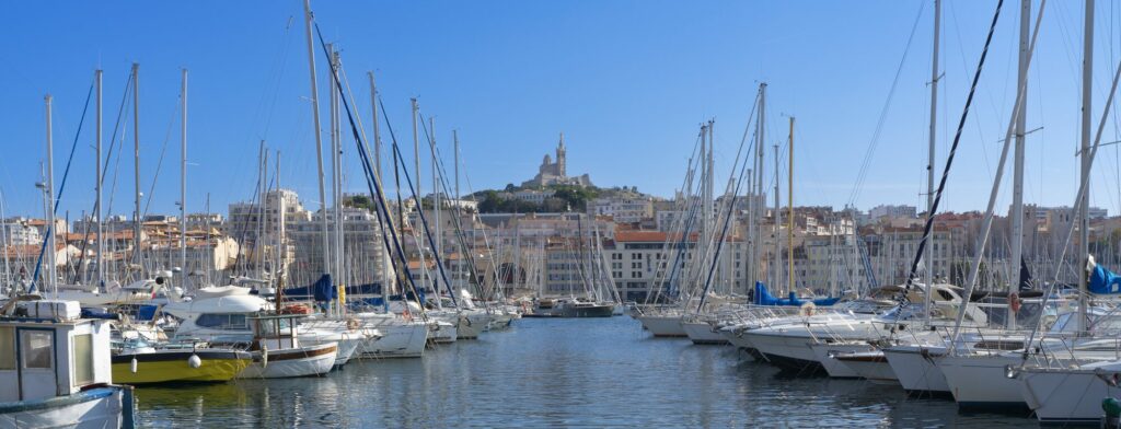 Voiliers amarrés dans le Vieux-Port de Marseille sous un ciel bleu, avec Notre-Dame-de-la-Garde sur la colline au loin