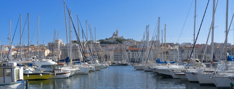 Voiliers amarrés dans le Vieux-Port de Marseille sous un ciel bleu, avec Notre-Dame-de-la-Garde sur la colline au loin