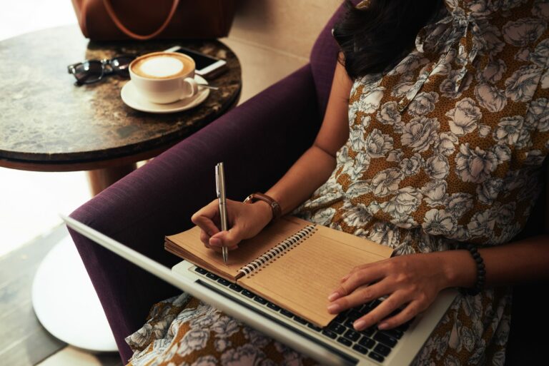 Femme écrivant dans un carnet à une table de café, à côté d’un ordinateur portable et d’une tasse