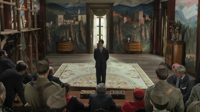 Un jeune homme en costume sombre debout au centre d'un salon aux murs peints, face à une assemblée de militaires et de notables, dans le film La Bola Negra