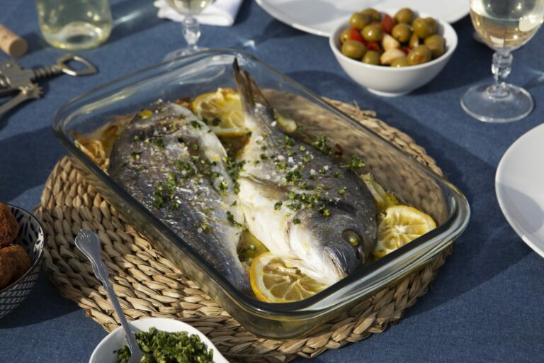 Deux daurades entières rôties aux herbes et au citron dans un plat en verre, sur une table au soleil dressée d'une nappe bleue avec verres de vin blanc et olives