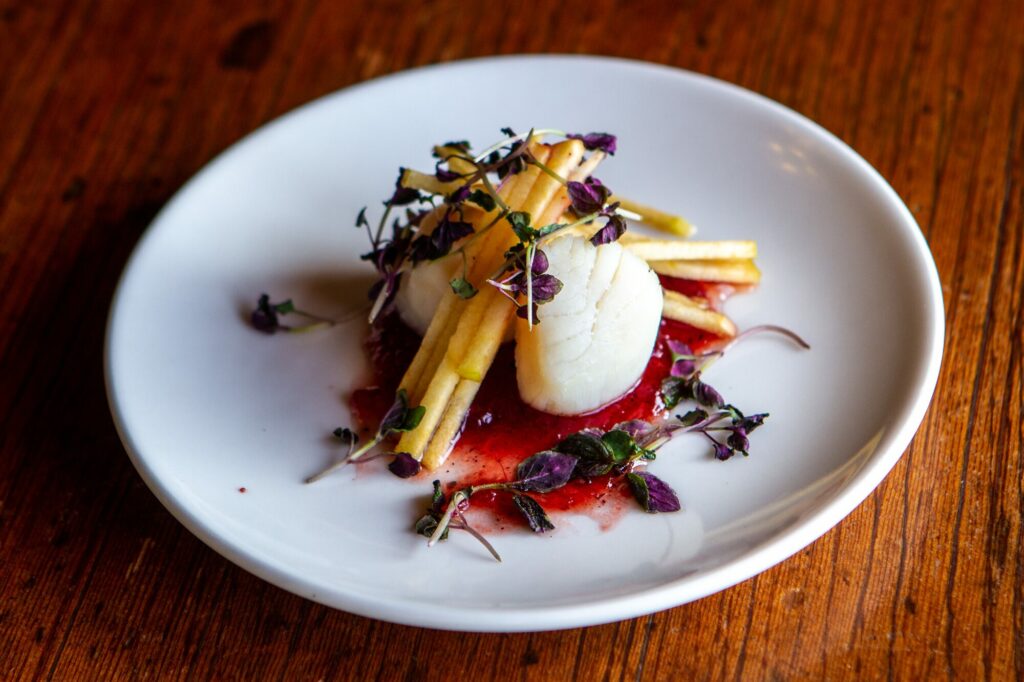 Assiette gastronomique de saint-jacques et de légumes dressée sur une assiette blanche posée sur une table en bois