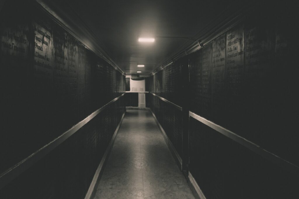 Long couloir sombre et étroit faiblement éclairé par des lampes au plafond, avec une porte claire tout au fond