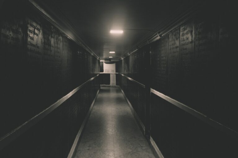 Long couloir sombre et étroit faiblement éclairé par des lampes au plafond, avec une porte claire tout au fond