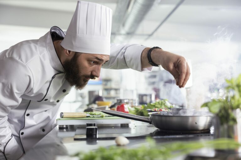 Un chef barbu en veste blanche et toque dresse une assiette d’herbes fraîches dans une cuisine professionnelle
