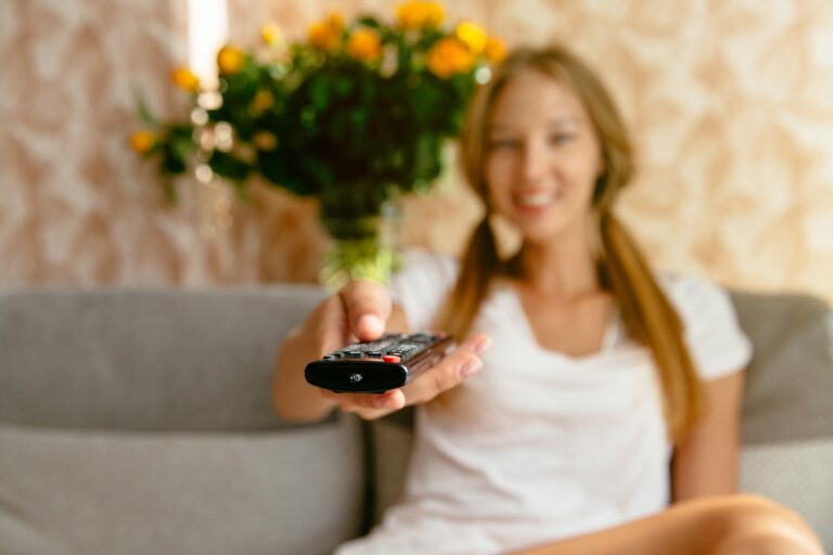 Une jeune femme assise sur un canapé pointe une télécommande vers la télévision, devant un bouquet de fleurs jaunes