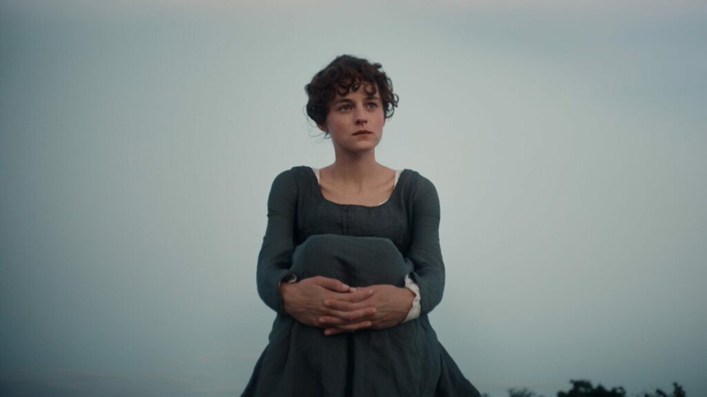 Emma Corrin en Elizabeth Bennet, en robe d’époque verte, debout dans un champ sous un ciel nuageux dans la série Orgueil et Préjugés