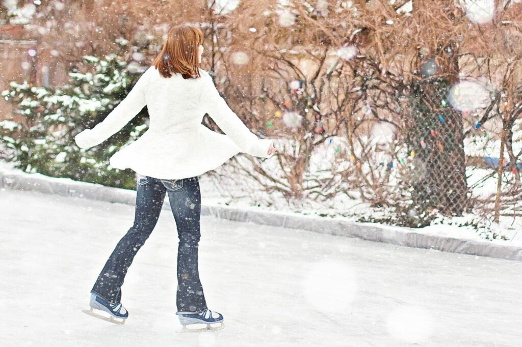 Patineuse en manteau blanc, vue de dos, glissant sur une patinoire en plein air bordée d’arbres enneigés