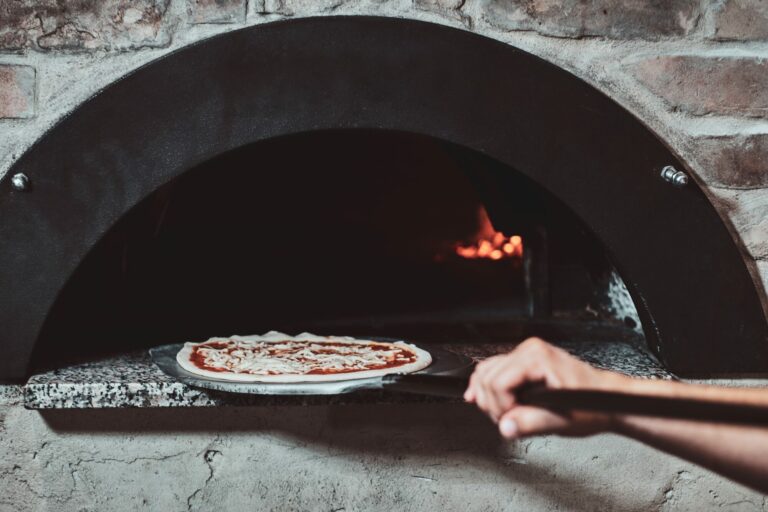 Pizza margherita enfournée sur une pelle dans un four à bois en briques où brûle une flamme