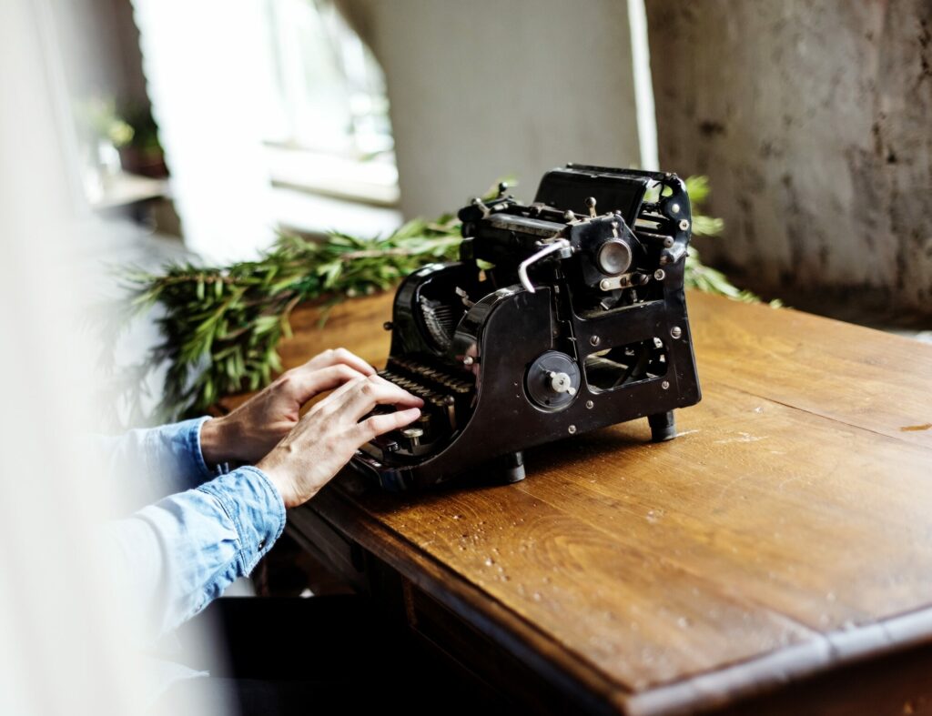 Mains tapant sur une machine à écrire ancienne posée sur une table en bois, près d’une fenêtre