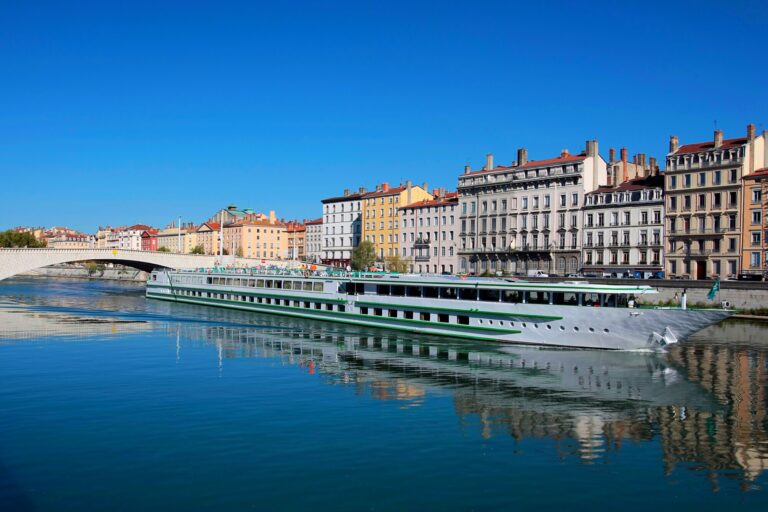 Les quais de la Saône à Lyon sous un ciel bleu, avec un bateau de croisière amarré devant une rangée d'immeubles anciens et un pont en arrière-plan