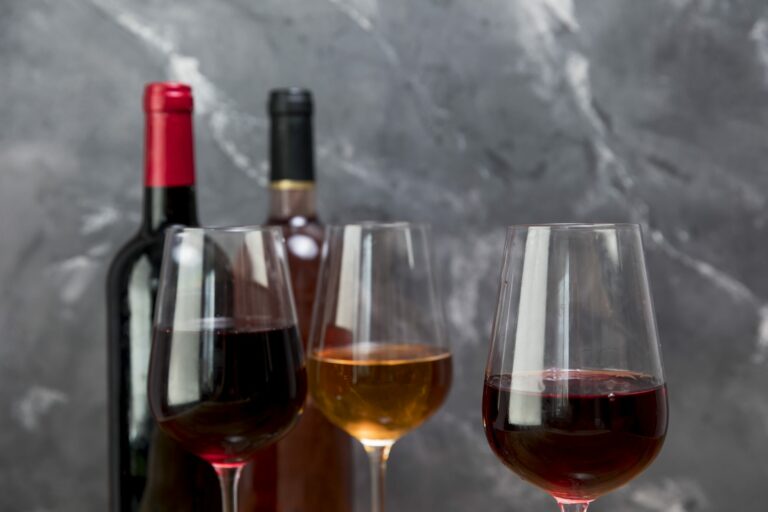 Trois verres de vin rouge, rosé et rouge devant deux bouteilles sur un fond de marbre gris