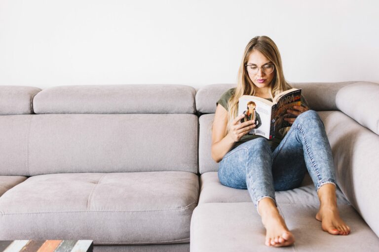 Jeune femme pieds nus assise sur un canapé gris, plongée dans la lecture d'un livre