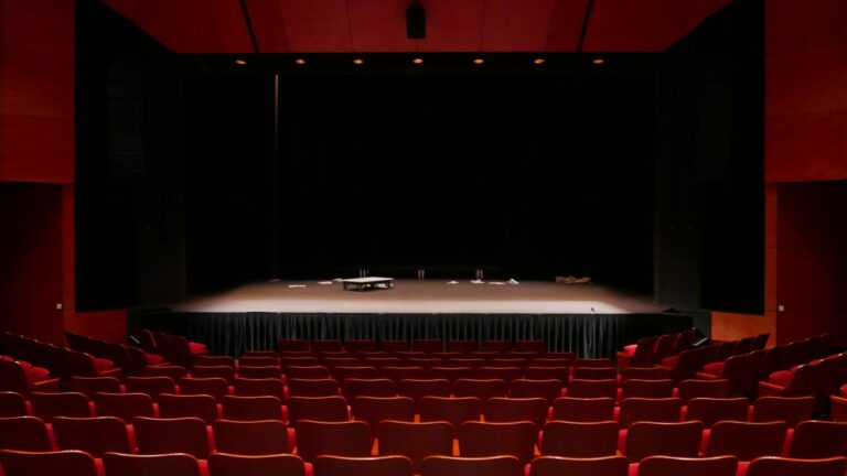 Salle de spectacle vide aux fauteuils et murs rouges, face à une grande scène encadrée de rideaux noirs
