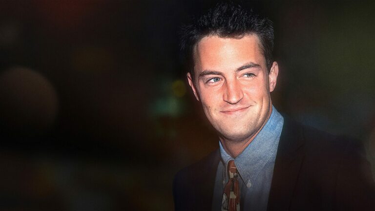 Matthew Perry souriant, en costume sombre, chemise bleue et cravate à motifs, sur un fond sombre