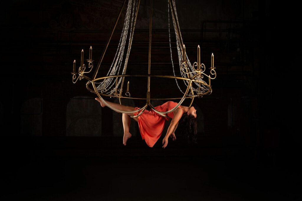 Acrobate en tenue rouge suspendue sous un cerceau aérien orné de guirlandes lumineuses, sur fond noir
