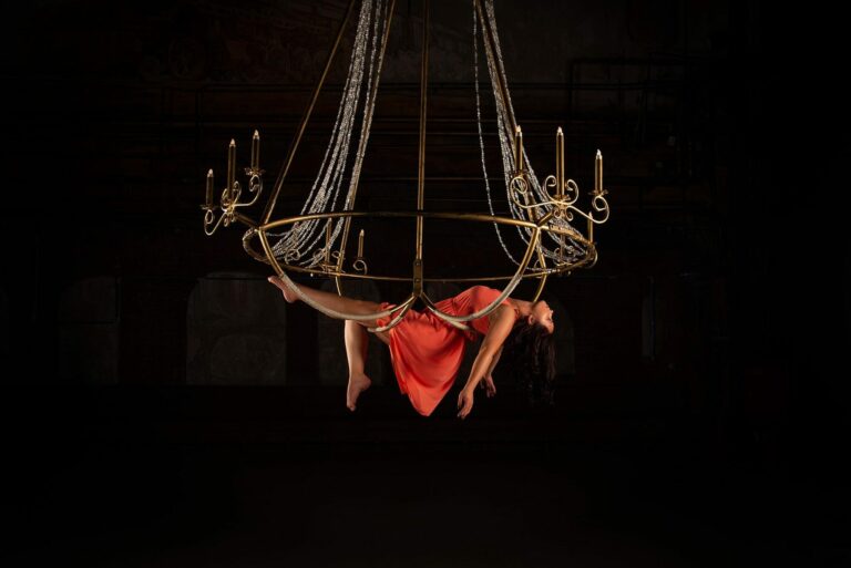 Acrobate en tenue rouge suspendue sous un cerceau aérien orné de guirlandes lumineuses, sur fond noir