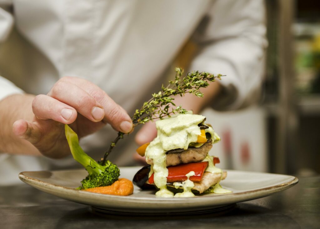 Mains d'un chef en veste blanche déposant une herbe fraîche sur un plat gastronomique dressé en hauteur